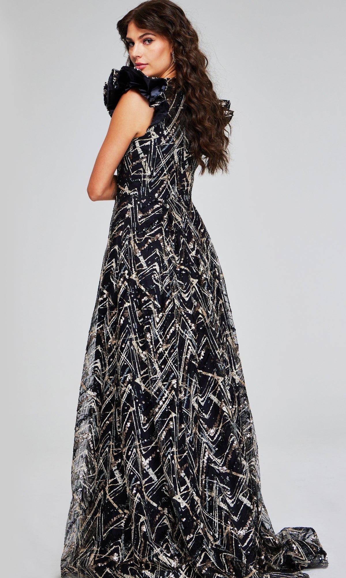 A woman with long wavy hair poses with her back to the camera in a glamorous Jovani 39274 long formal dress, featuring a sleeveless design and a floor-length black and silver pattern.